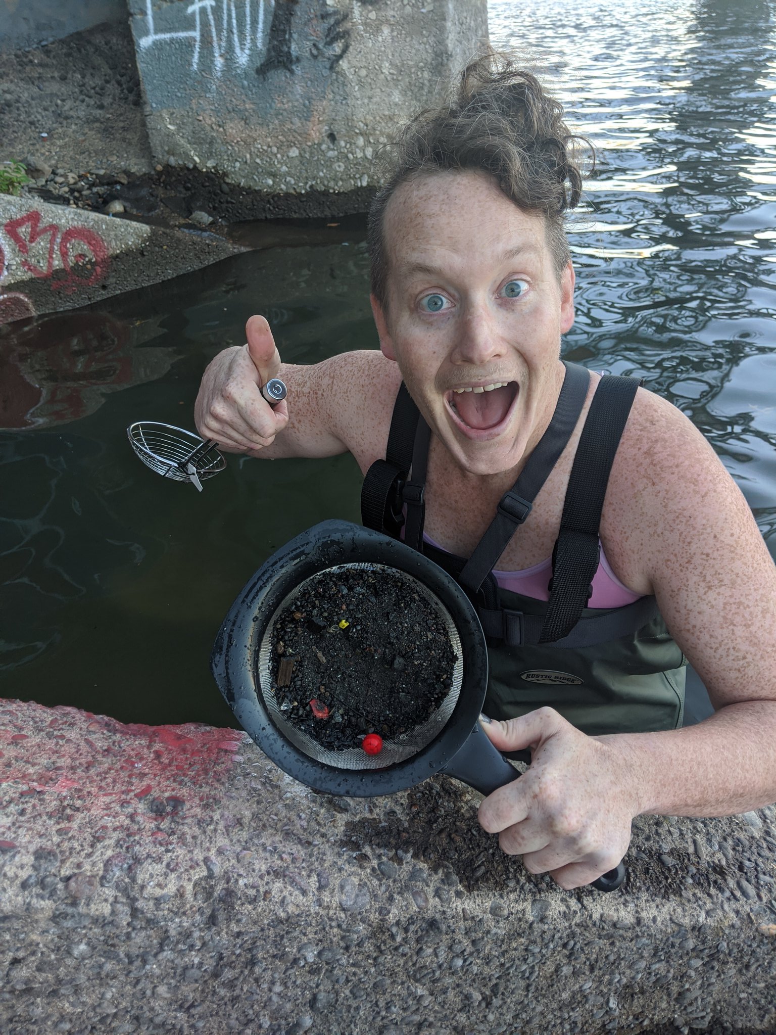 Juniper standing in water in waders giving a thumbs up and holding a sieve with sediment from the Willamette River, including an intact Pepperball.